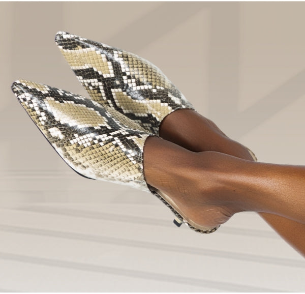 A pair of Kelley Kouture Debut pointed-toe kitten heel mules with a beige, brown, and black snakeskin print pattern, shown on a model's feet against a neutral light beige background.