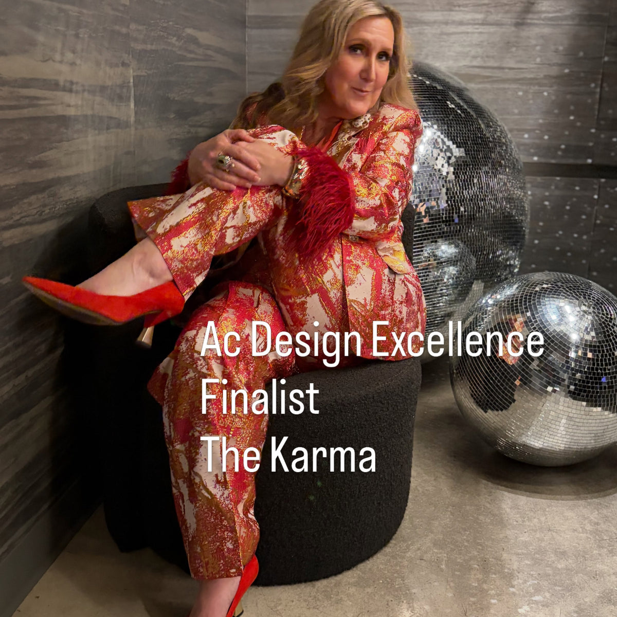Kelley Williamson sits on a black round ottoman in a styled interior, wearing a red and gold patterned fringed jacket and matching pants from Ukay's Closet with a pair of Kelley Kouture Karma Signature Pumps in red. Large mirrored disco balls and a gray wood-paneled wall are behind her, with white text overlay reading 'Ac Design Excellence Finalist The Karma'.