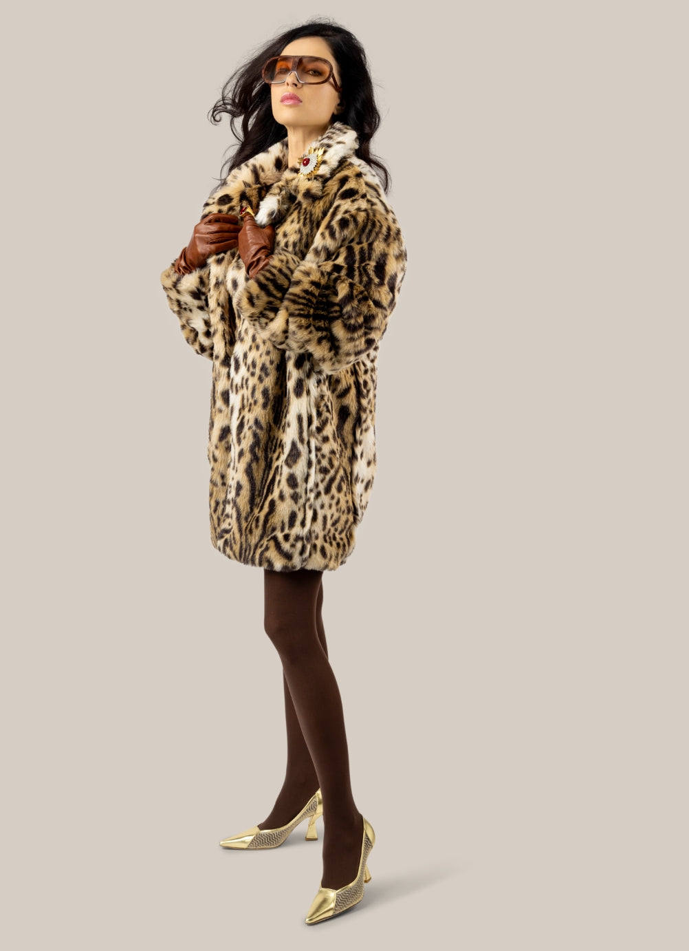 A woman modeling a leopard-print faux fur coat, styled with brown tights, wearing Kismet gold metallic napa with gold glitter on nude mesh signature Kelley Kouture two-piece K heels, and sunglasses, against a solid light gray background.