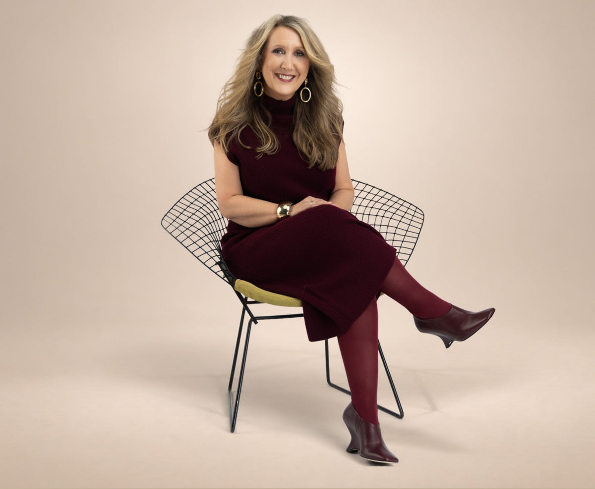 Kelley Williamson sits on a modern wire chair against a warm beige studio background, wearing a dark burgundy sleeveless dress, matching tights, and a pair of Kelley Kouture Ellie booties in Black Cherry.