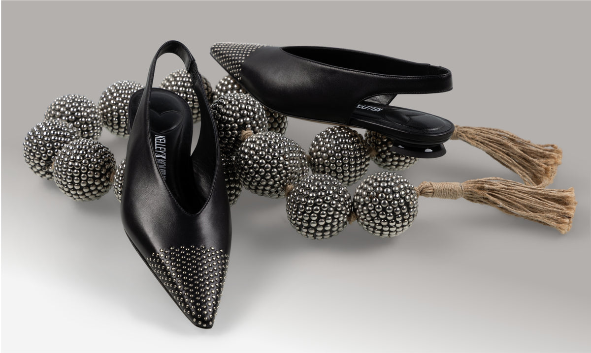 A pair of Kelley Kouture Luna in black. Leather slingback flat pump with demi-wedge heel, arranged on a neutral gray background with decorative rhinestone spheres and tassel garlands.